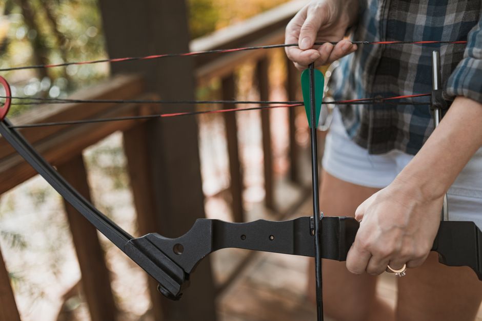 A person holding a black bow and an arrow, preparing to shoot outdoors on a wooden deck or balcony. The individual is wearing a plaid shirt and white shorts, with sunlight filtering through the trees 